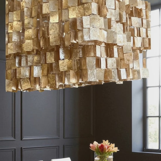 A unique capiz shell chandelier with layered, translucent champaign/golden tiles hangs above a dark colored dining table, casting a warm, soft glow. Below, a simple floral arrangement sits at the center, while dark gray wainscoting on the walls and a nearby window create contrast. The textured fixture adds elegance and visual interest, creating a modern yet organic atmosphere.  A focal point for any living room, kitchen, dining area, or foyer.  