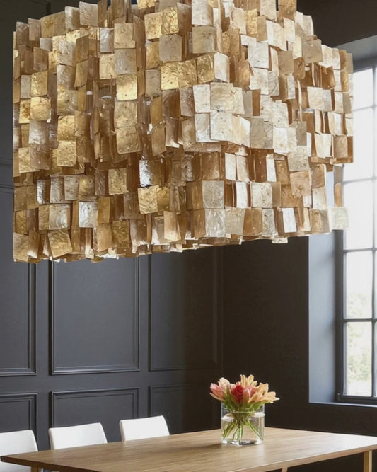 A unique capiz shell chandelier with layered, translucent champaign/golden tiles hangs above a dark colored dining table, casting a warm, soft glow. Below, a simple floral arrangement sits at the center, while dark gray wainscoting on the walls and a nearby window create contrast. The textured fixture adds elegance and visual interest, creating a modern yet organic atmosphere.  A focal point for any living room, kitchen, dining area, or foyer.  