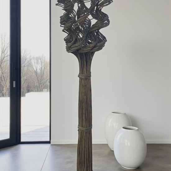 A tall floor-standing wicker sculpture made of bundled natural fibers stands in a minimalist room. The base is tightly bound, extending upward into an intricate, twisted woven design that creates an abstract, sculptural form. It is placed near a large window with natural light, with two smooth white ceramic floor vases in the background, highlighting its organic texture and vertical presence.