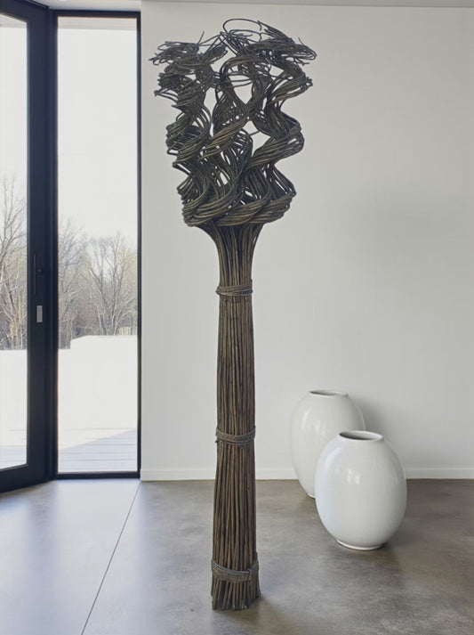 A tall floor-standing wicker sculpture made of bundled natural fibers stands in a minimalist room. The base is tightly bound, extending upward into an intricate, twisted woven design that creates an abstract, sculptural form. It is placed near a large window with natural light, with two smooth white ceramic floor vases in the background, highlighting its organic texture and vertical presence.