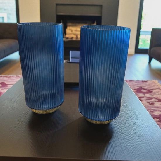 Two matching blue fluted glass vases with a ribbed texture sit on a dark coffee table in a modern living room. The deep blue color and vertical lines create a sleek, elegant look. Sofas, a fireplace, and a patterned rug are visible in the background, adding warmth and context to the contemporary setting.