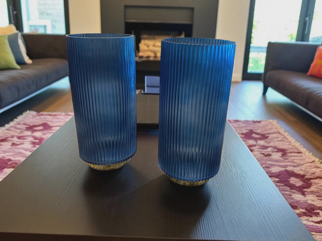 Two matching blue fluted glass vases with a ribbed texture sit on a dark coffee table in a modern living room. The deep blue color and vertical lines create a sleek, elegant look. Sofas, a fireplace, and a patterned rug are visible in the background, adding warmth and context to the contemporary setting.