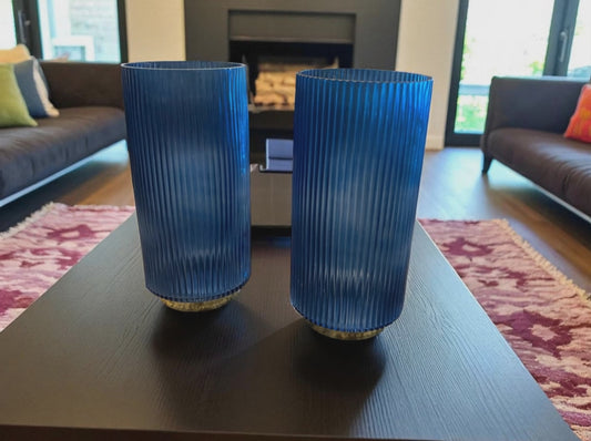 Two matching blue fluted glass vases with a ribbed texture sit on a dark coffee table in a modern living room. The deep blue color and vertical lines create a sleek, elegant look. Sofas, a fireplace, and a patterned rug are visible in the background, adding warmth and context to the contemporary setting.
