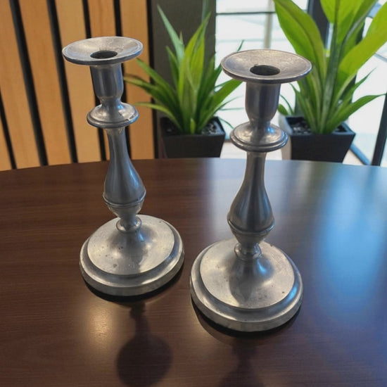 Two metal candle holders with a classic, sculptural design and aged silver finish sit on a dark wooden table. Behind them, green potted plants and a large window,  add a natural touch, while soft light highlights their smooth curves and subtle patina. The scene creates a warm, elegant, and timeless atmosphere with a blend of natural and refined elements.