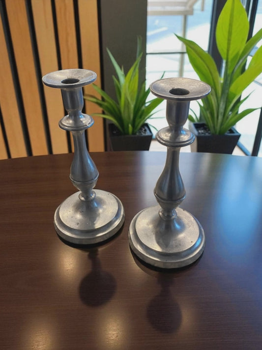 Two metal candle holders with a classic, sculptural design and aged silver finish sit on a dark wooden table. Behind them, green potted plants and a large window,  add a natural touch, while soft light highlights their smooth curves and subtle patina. The scene creates a warm, elegant, and timeless atmosphere with a blend of natural and refined elements.